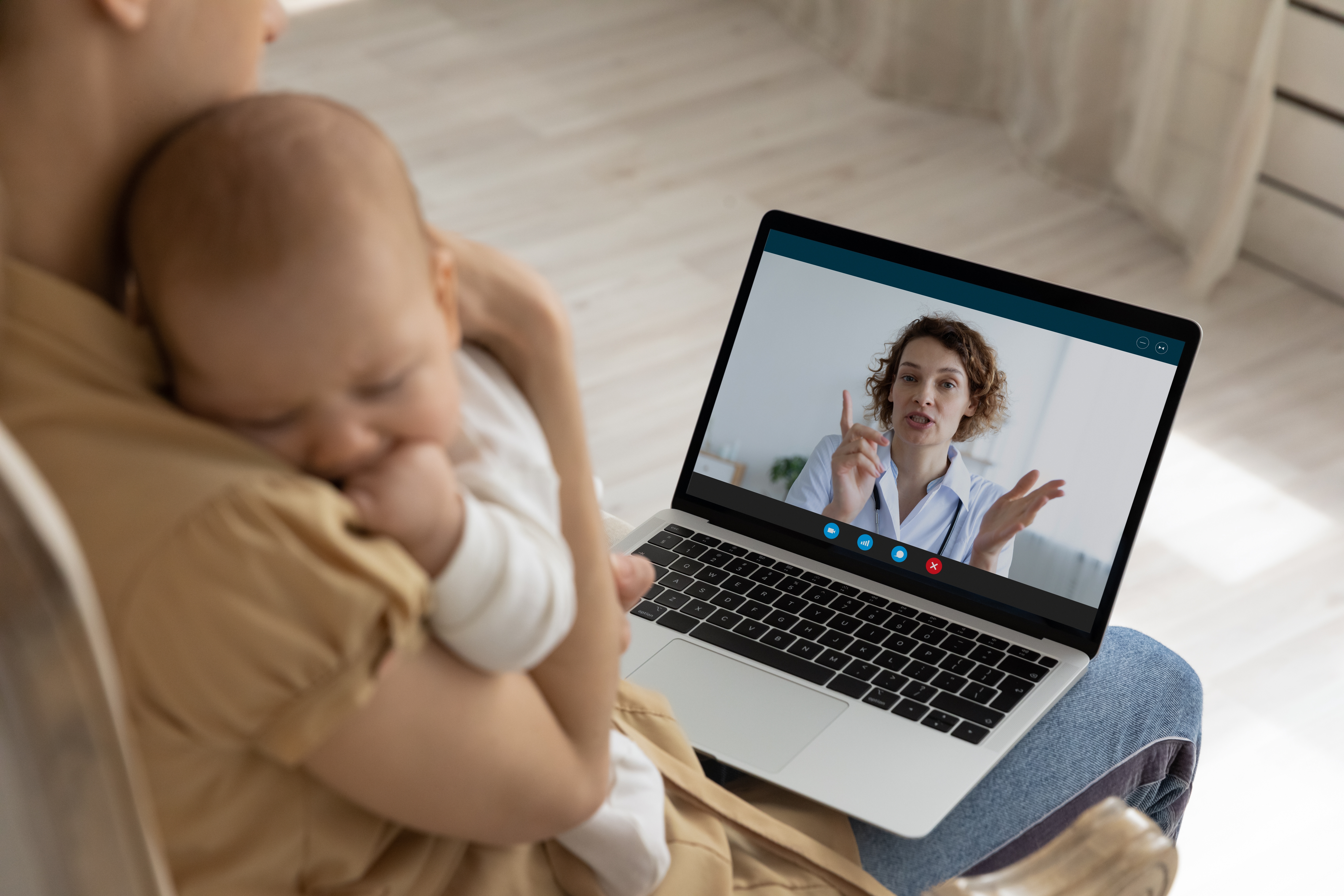 A mother holds a baby while talking to her doctor using a video telehealth appointment on her laptop.