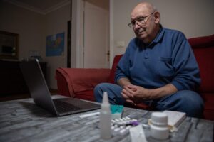 A man sits in front of his laptop having a video telehealth appointment. His medications are sitting on the table beside the laptop.
